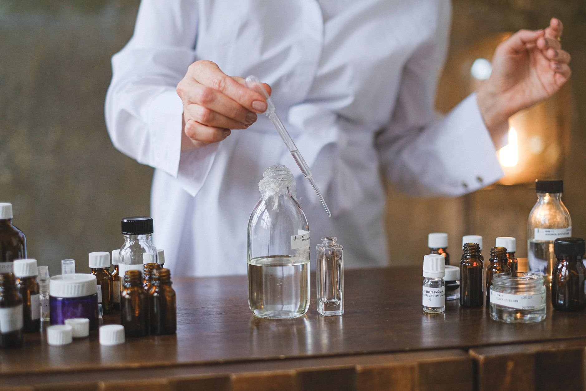 Personne en blouse de laboratoire utilisant une pipette pour transférer du liquide dans des bouteilles en verre transparent sur une table.