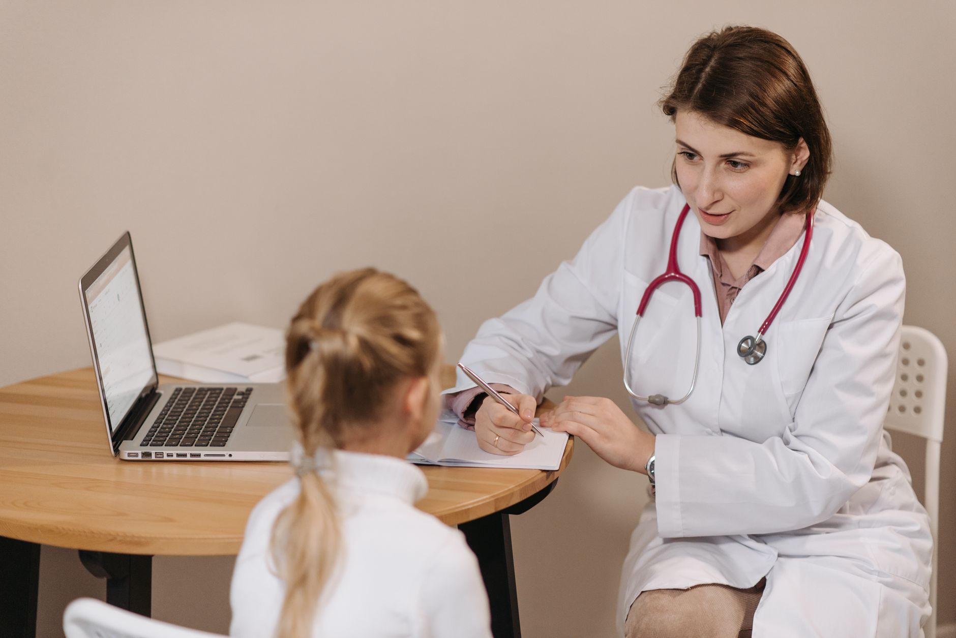 Une médecin en blouse blanche écrit des notes à une table en bois face à une jeune patiente vue de dos, avec un ordinateur portable ouvert.
